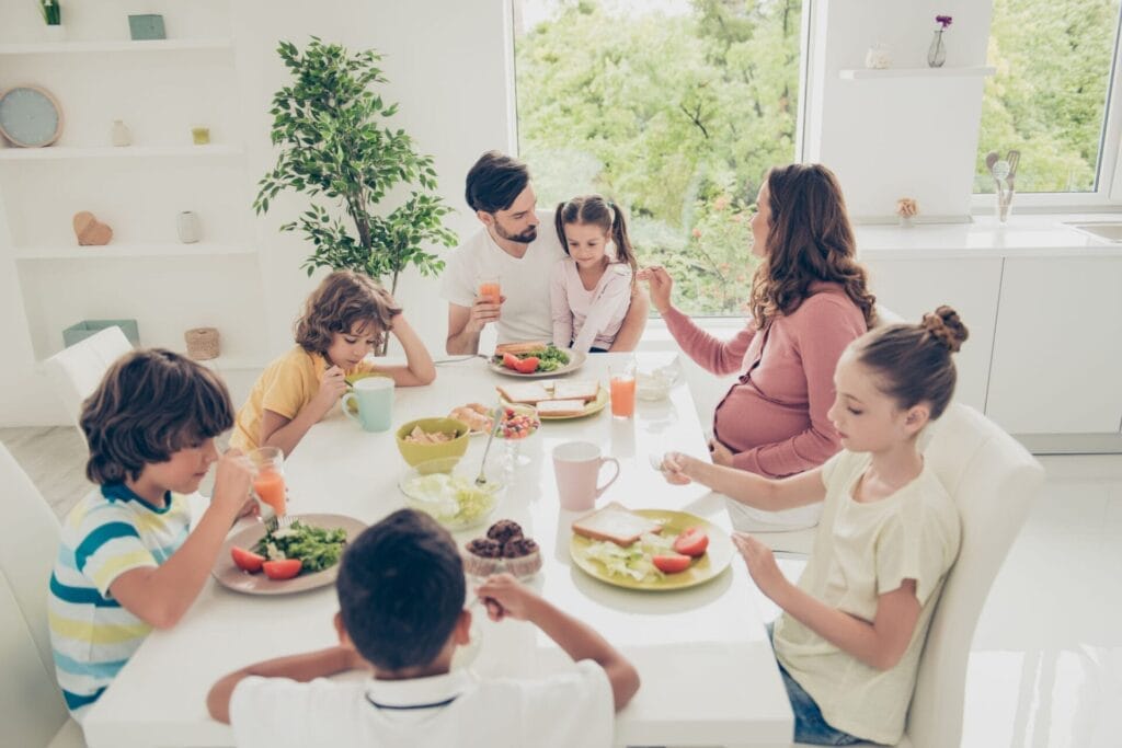 Big nice adorable beautiful lovely family, mulato brothers and sisters having dinner, lunch, eating crispy toasts, salad, vegs, girl on father's hands, mom feeding girl, white light interior dining